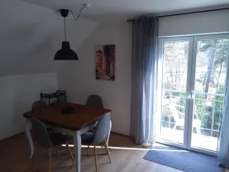 Ein gemütlicher Essbereich mit einem Holztablett und grauen Stühlen. Große Fenster lassen viel Tageslicht herein und bieten einen Ausblick ins Freie.