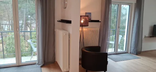 Ein gemütlicher Wohnbereich mit einem schwarzen Sessel und warmem Licht. Große Fenster bieten Blick auf die Natur draußen.