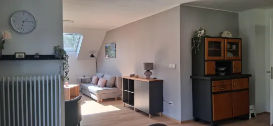 Ein helles Wohnzimmer mit einer gemütlichen Couch und einem modernen Regal. An der Wand hängt eine Uhr, und es gibt einen kleinen Esstisch mit Stühlen.