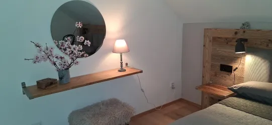 Ein gemütliches Schlafzimmer mit einem Holzkopfteil und einer Tischlampe. Eine dekorative Vase mit Blüten und ein runder Spiegel sorgen für einen modernen Look.