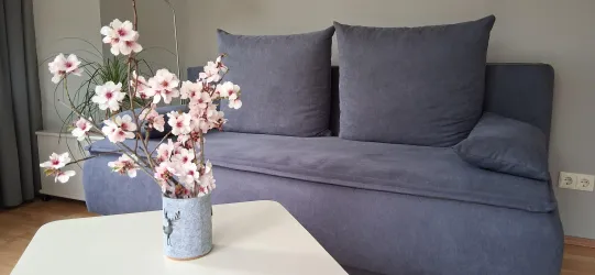 Ein modernes Sofa mit großen Kissen steht in einem gemütlichen Raum. Auf einem Tisch daneben befindet sich eine Vase mit blühenden Zweigen.
