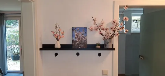Eine helle Wand mit einem schwarzen Regal, auf dem zwei Vasen mit Blumen und ein Bild stehen. Daneben sind Haken an der Wand angebracht.