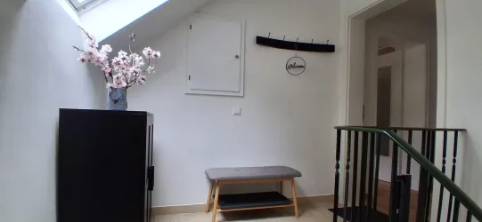 Ein heller Flur mit einer Bank und einem schwarzen Schrank. Auf der Bank steht eine Vase mit künstlichen Blumen und an der Wand hängt ein Schild mit der Aufschrift "Willkommen".