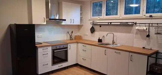 Eine moderne Küche mit weißen Schränken und Holzoberflächen. Das Bild zeigt einen Herd, einen Kühlschrank und große Fenster, die viel Licht hereinlassen.