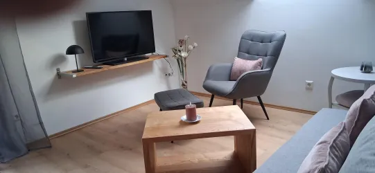 Ein gemütliches Wohnzimmer mit einem Fernseher an der Wand und einem modernen Sessel. Auf dem Couchtisch steht eine Kerze und eine kleine Blumenvase.