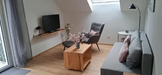Ein modernes Wohnzimmer mit einem Fernseher, einem grauen Sofa und einem Holzkaffeetisch. Große Fenster sorgen für viel Tageslicht und eine einladende Atmosphäre.