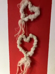 Zwei weiße Herzen aus Wolle hängen an einer roten Wand. Sie sind kunstvoll miteinander verbunden und wirken freundlich und einladend.