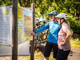 Zwei Personen befinden sich mit ihrem Mountainbike am Haus der Nachhaltigkeit in Johanniskreuz und schauen sich eine Wegekarte an.