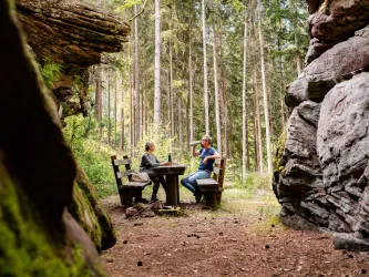 Zwei Personen sitzen an einem Rastplatz aus zwei Holzbänken und einem Holztisch, der zwischen dem Nibelungenfelsen liegt. Sie trinken Wasser und rasten.