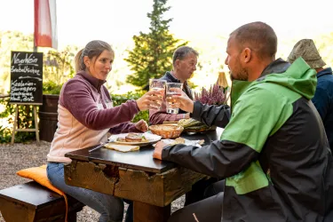 Zwei Personen stoßen mit dem Schorleglas bei einer Pfälzerwaldhütte an. Auf dem Tisch stehen typisch pfälzische Gerichte.  