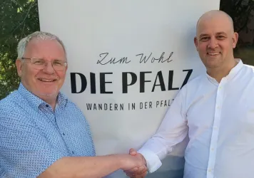 Hans-Ulrich Ihlenfeld und Martin Brandl schütteln sich die Hand vor einem Banner mit dem Zum Wohl die Pfalz Logo darauf.