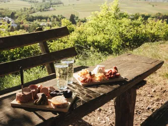 Ein Rastplatz aus Holz in der Natur. Auf dem Tisch stehen zum Wohl die Pfalz Dubbegläser und Vesperplatten mit Pfälzischer Brotzeit.