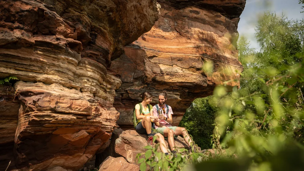 Ein Paar sitzt vor einer Sandsteinfelswand im Pfälzerwald und picknickt.