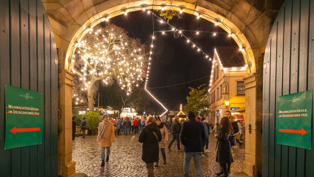 Besuchende des Deidesheimer Weihnachtsmarkt schlendern entlang der Buden durch das Winzerdorf.