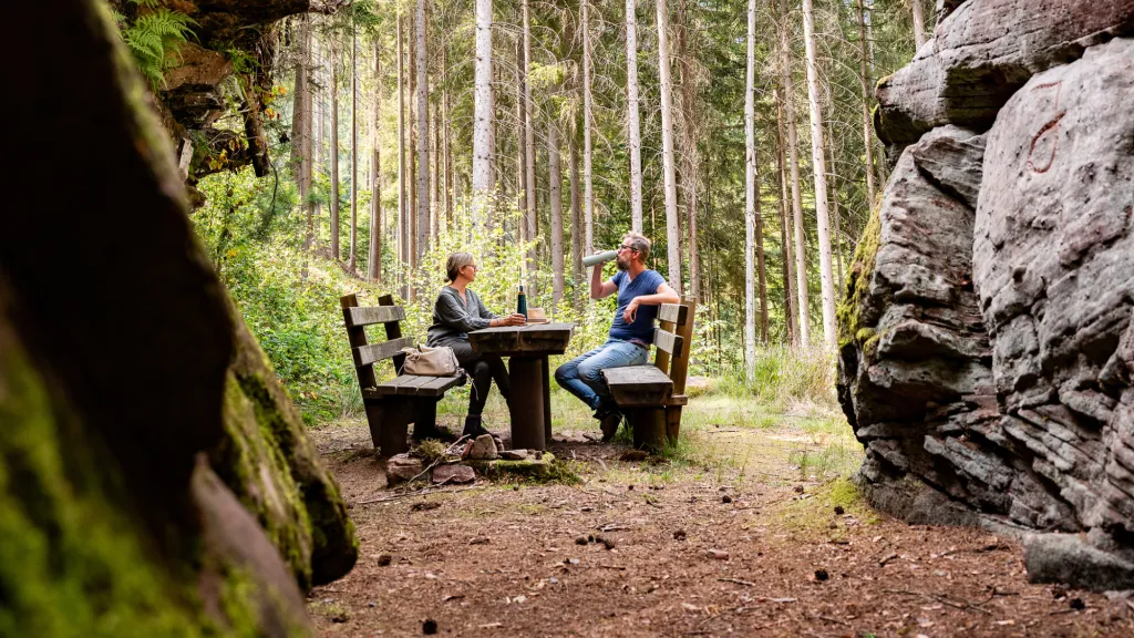Zwei Personen sitzen an einem Rastplatz aus zwei Holzbänken und einem Holztisch, der zwischen dem Nibelungenfelsen liegt. Sie trinken Wasser und rasten.