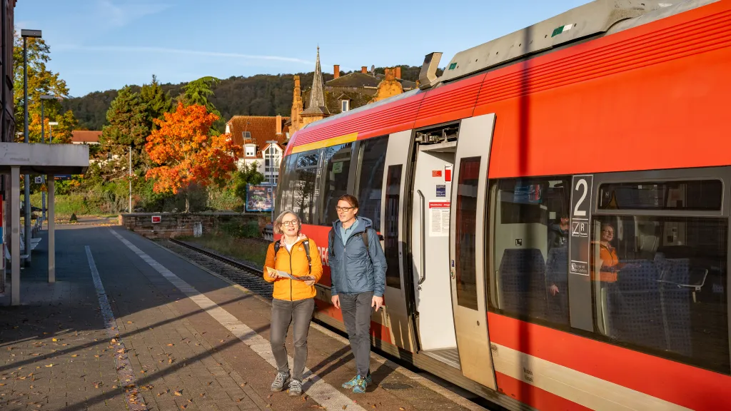 Zwei Personen steigen aus einem Zug. Sie tragen Wanderkleidungen und machen sich auf den Weg zu einer Wanderung in der Pfalz.