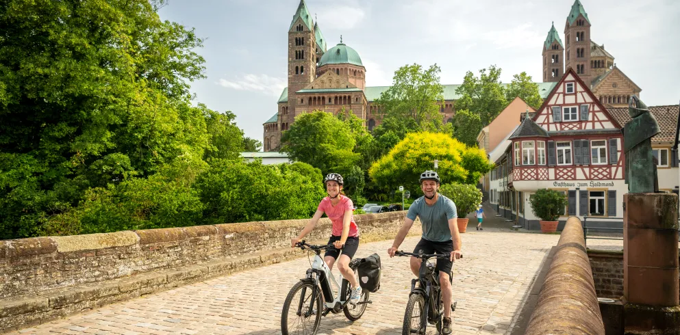 Zwei Personen fahren mit dem Rad über die Sonnenbrücke in Speyer. Im Hintergrund ist der Dom und die Fachwerkhäuser.