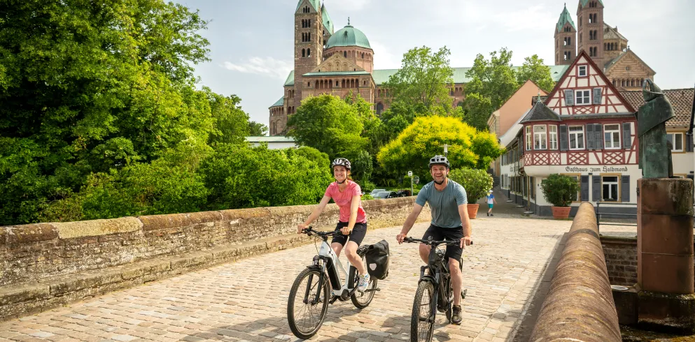 Zwei Personen fahren mit dem Rad über die Sonnenbrücke in Speyer. Im Hintergrund ist der Dom und die Fachwerkhäuser.