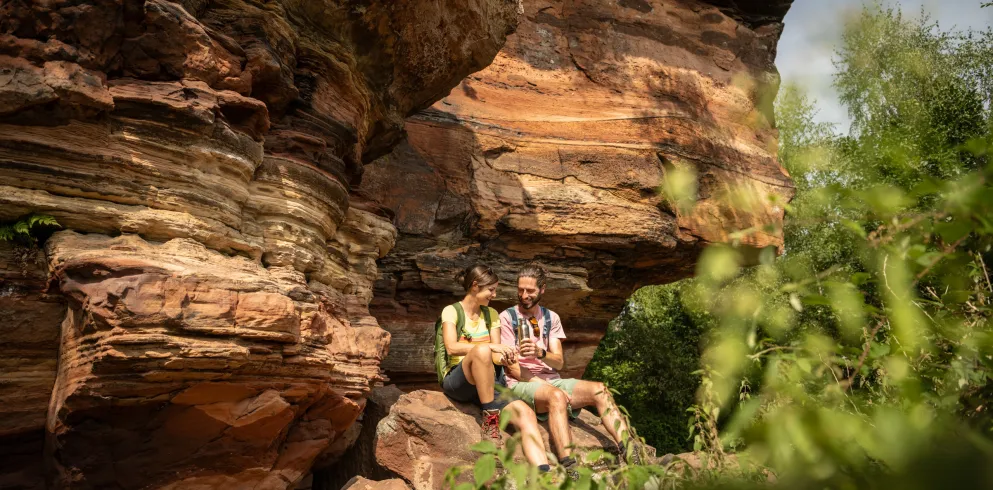 Ein Paar sitzt vor einer Sandsteinfelswand im Pfälzerwald und picknickt.