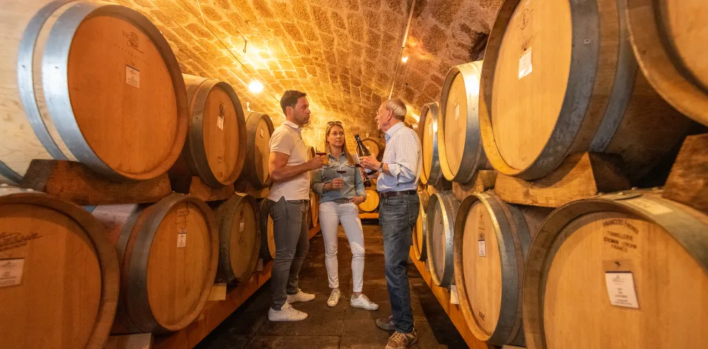 In einem Weinkeller stehen zwei Personen vor großen Weinfässern und kosten die Weine, die ein Winzer präsentiert. 