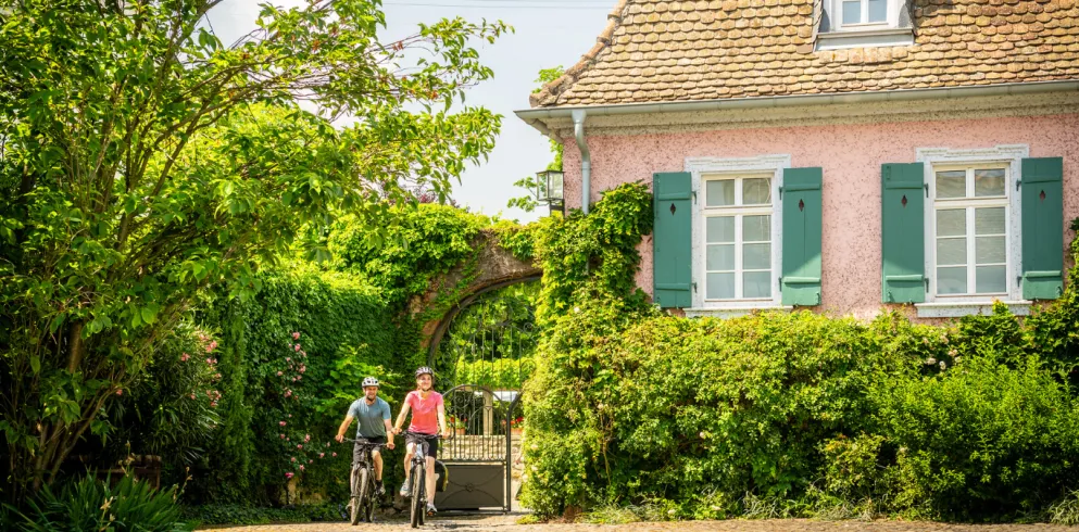 Zwei Personen Fahren mit dem Rad an einem alten rosafarbenen Haus vorbei.
