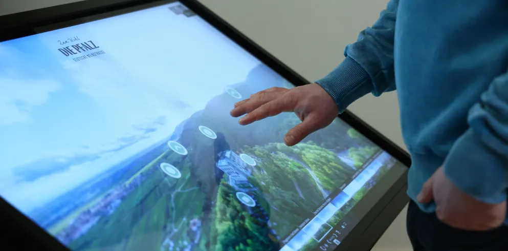 ine Person bedient eine digitale Stele, auf deren Bildschirm eine Luftaufnahme der Pfalz ist.