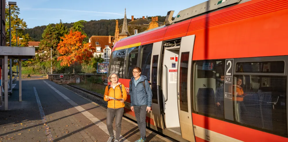 Zwei Personen steigen aus einem Zug. Sie tragen Wanderkleidungen und machen sich auf den Weg zu einer Wanderung in der Pfalz.