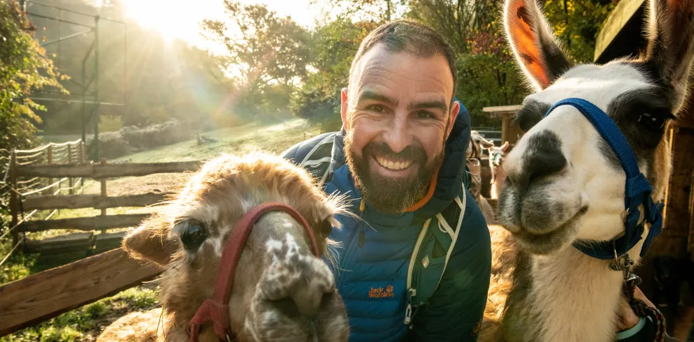 Eine Person lächelt in eine Kamera. Neben der Person stehen zwei Lamas.