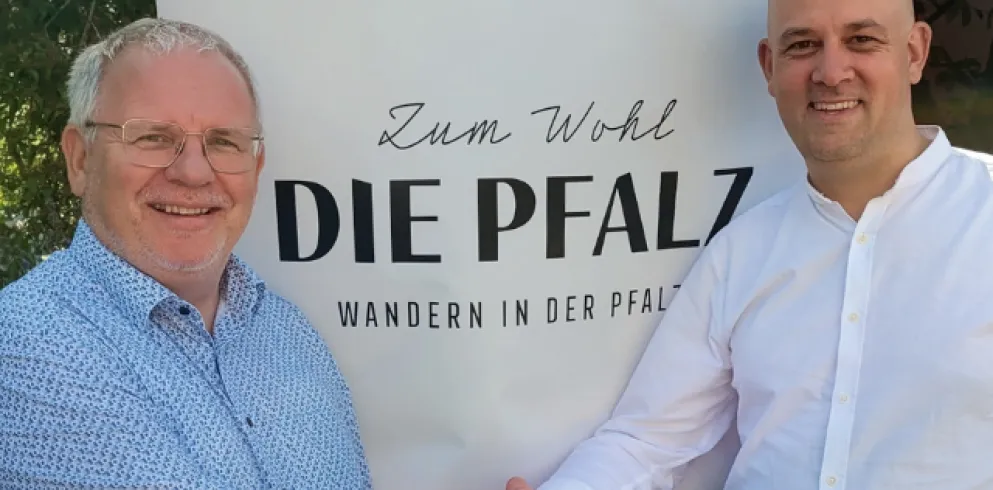 Hans-Ulrich Ihlenfeld und Martin Brandl schütteln sich die Hand vor einem Banner mit dem Zum Wohl die Pfalz Logo darauf.