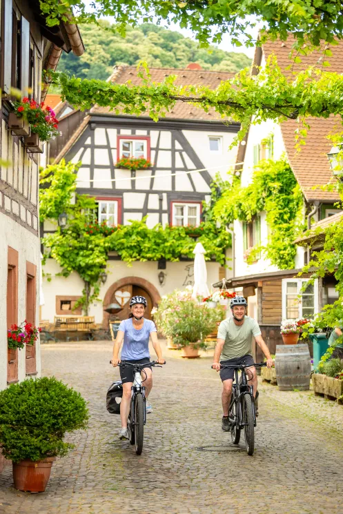 Zwei Personen mit Rad fahren entlang Fachwerkhäuser in einem Winzerdorf an der Deutschen Weinstraße