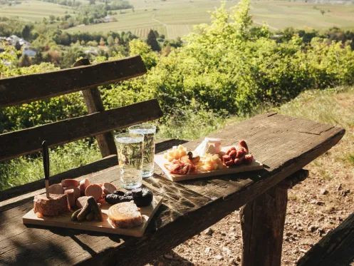 Ein Rastplatz aus Holz in der Natur. Auf dem Tisch stehen zum Wohl die Pfalz Dubbegläser und Vesperplatten mit Pfälzischer Brotzeit.
