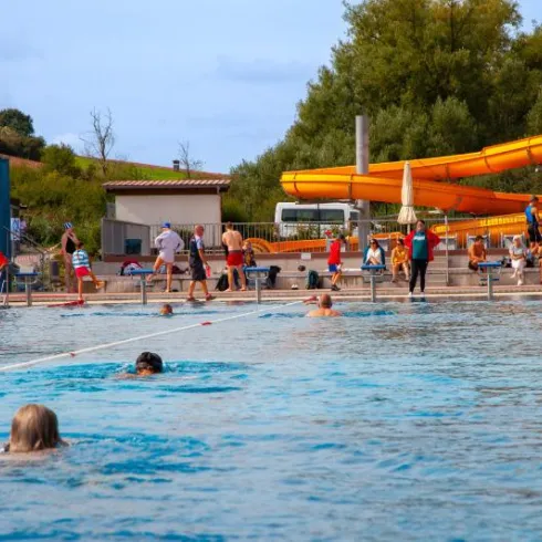 Ein Schwimmbecken mit vielen Menschen, die schwimmen und sich entspannen. Im Hintergrund sind Rutschen und Sonnenschirme zu sehen.