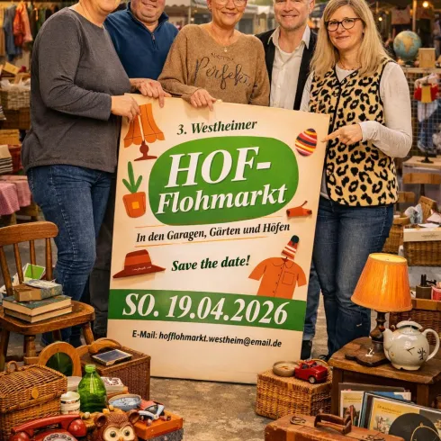 Fünf Personen stehen lächelnd vor einem Schild für den Hof-Flohmarkt. Im Hintergrund sind Stände und verschiedene Gegenstände zu sehen.