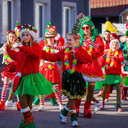 Eine fröhliche Parade mit verkleideten Teilnehmern in bunten, festlichen Outfits. Die Menschen tragen Weihnachtsdekorationen und tanzen in einer lebhaften Atmosphäre.