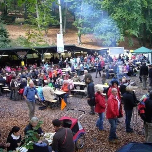 Eine lebhafte Veranstaltung im Wald mit vielen Menschen, die an Holztischen essen und Getränke genießen. Im Hintergrund sieht man Stände und eine gemütliche Atmosphäre.