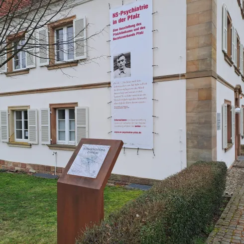 Ein historisches Gebäude mit einer Informationstafel im Vordergrund. An der Wand hängt ein großes Plakat mit Informationen zur Psychiatrie.