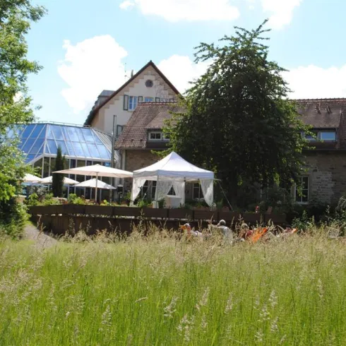 Ein charmantes Gebäude umgeben von grüner Wiesenlandschaft. Im Vordergrund stehen Sonnenschirme und Liegen für entspannte Stunden im Freien.