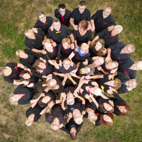 Eine Gruppe von Menschen steht im Kreis auf einer Wiese. Die Teilnehmer tragen schwarze Kleidung und zeigen verschiedene Gesten.