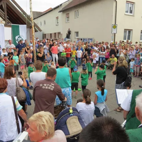 Eine lebhafte Versammlung von Menschen auf einer Straße bei einem festlichen Anlass. Kinder in grünen T-Shirts treten auf, während die Menge zuschaut und applaudiert.