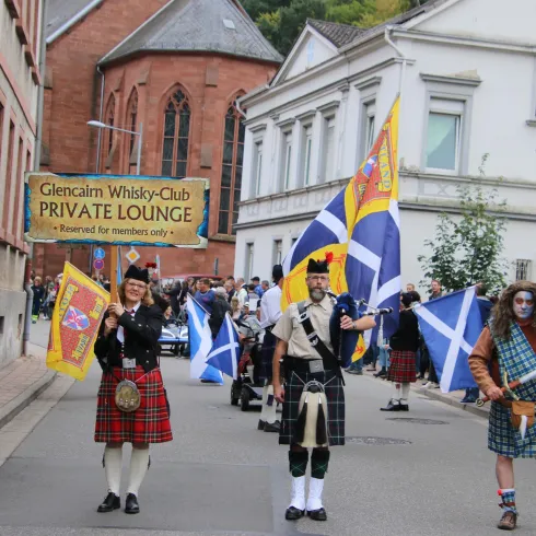 Eine Paraden mit Teilnehmern in traditionellen schottischen Outfits und Schottenröcken. Sie tragen Flaggen und ziehen durch eine belebte Straße.