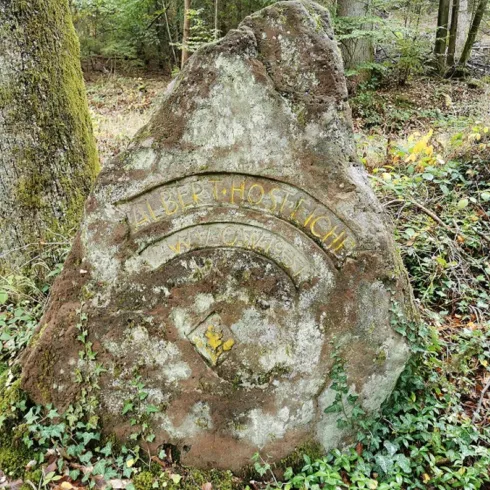 Ein großer, moosbedeckter Stein im Wald mit einer eingravierten Inschrift. Die Umgebung ist grün und naturbelassen.