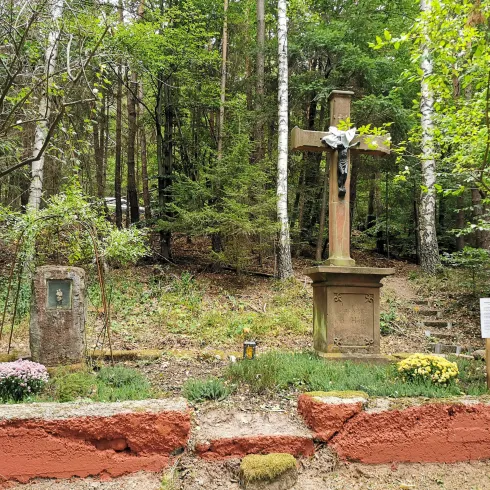 Ein ruhiger Gedenkplatz im Wald mit einem Kreuz und einer Stele. Umgeben von Bäumen und bunten Blumen.
