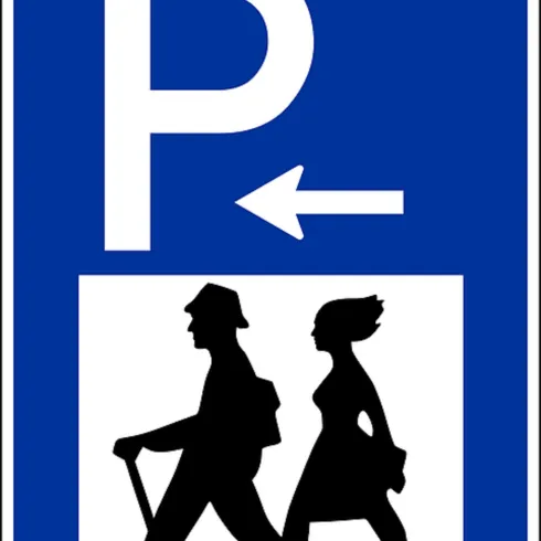 Ein Schild, das auf einen Parkplatz hinweist, mit einer pfeilartigen Richtung nach links. Darunter sind Silhouetten von zwei Personen, die spazieren gehen.