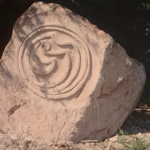 Ein großer, naturbelassener Stein mit einer kunstvollen Reliefdarstellung in der Mitte. Der Stein liegt auf dem Boden, umgeben von Gras und Büschen.