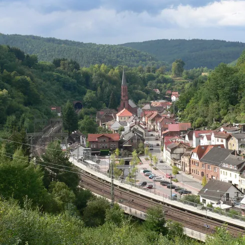 Eine kleines Dorf, Namens Weidenthal, im Tal mit einer Kirche und roten Dächern. Umgeben von grünen Hügeln und einer Eisenbahnlinie.