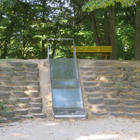 Eine einfache Rutsche in einem Park, umgeben von Bäumen. Im Hintergrund steht eine gelbe Bank.