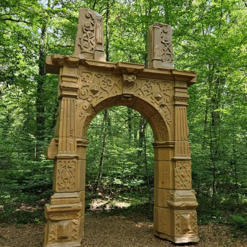 Ein kunstvoll gestalteter Torbogen aus Holz steht im Wald. Die umgebende Natur ist grün und einladend.