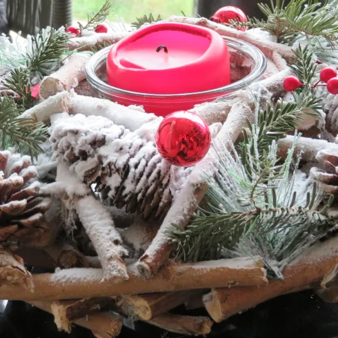 Ein festlicher Tischkranz aus Tannenzweigen, Tannenzapfen und roten Kugeln. In der Mitte steht eine pinke Kerze, umgeben von einem Hauch Schnee.