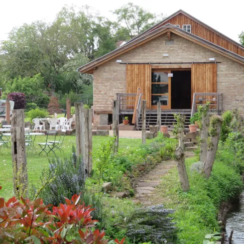 Ein charmantes Gebäude aus Stein mit Holzverkleidung, umgeben von grünem Gras und einem kleinen Bach. Im Vordergrund sieht man Sitzgelegenheiten und blühende Pflanzen.