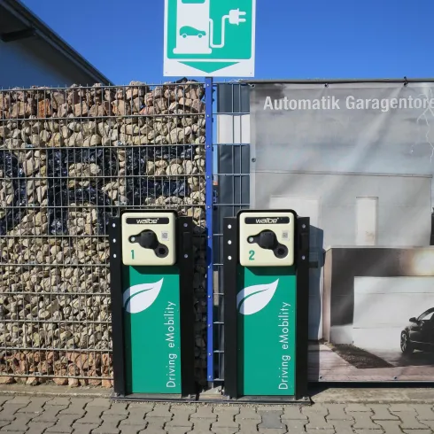 Zwei Elektroladestationen stehen nebeneinander an einer Wand. Darüber hängt ein Schild mit dem Symbol für Elektromobilität.